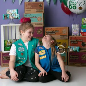 Two girl scouts sisters laughing candidly in front of their cookie packages.